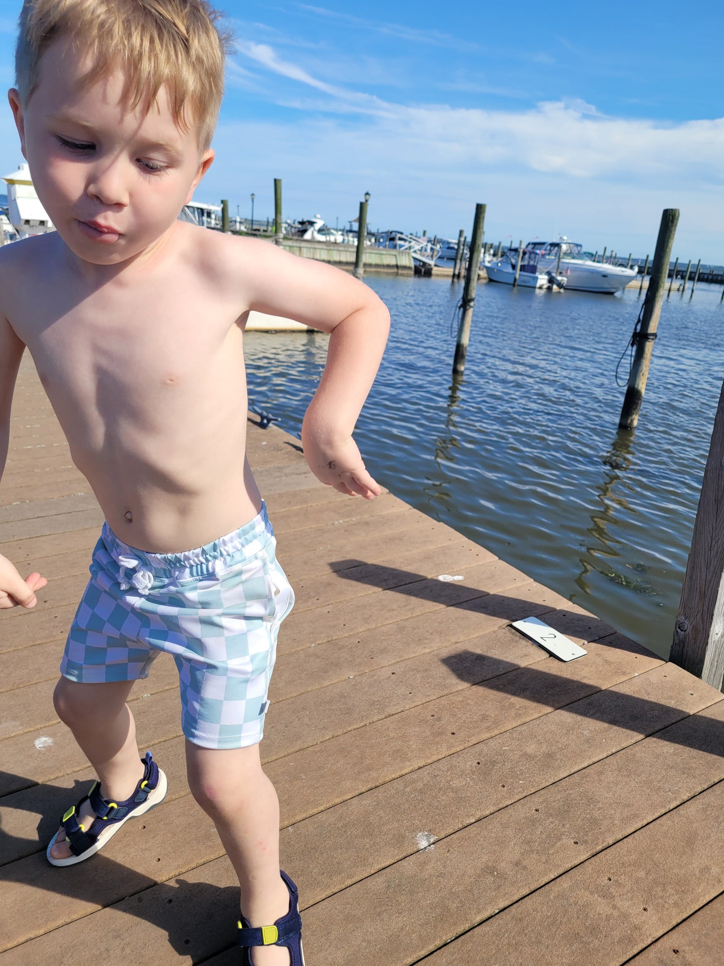 Toddler wearing Boys white and mint green checkered swim shorts, dancing on marina dock.