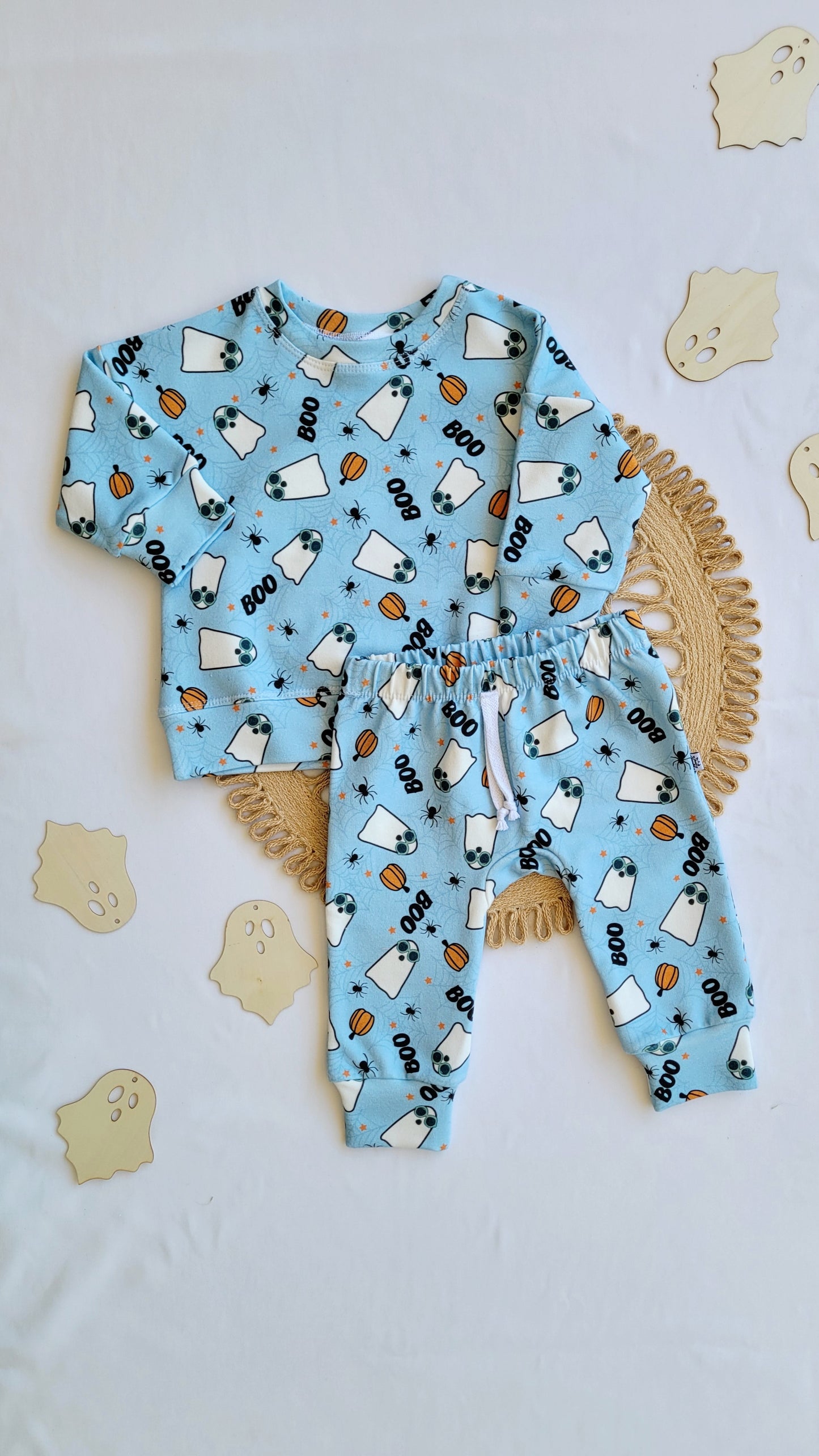 Flat lay of a two-piece toddler outfit featuring a light blue ghost-themed sweatshirt and matching sweatpants with Halloween designs, placed on a woven mat with wooden ghost decorations around.